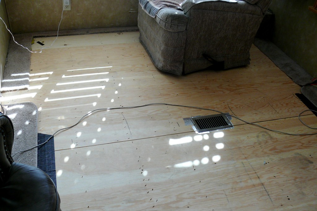 An RV interior with the carpet pulled up, exposing the plywood subfloor and a floor vent