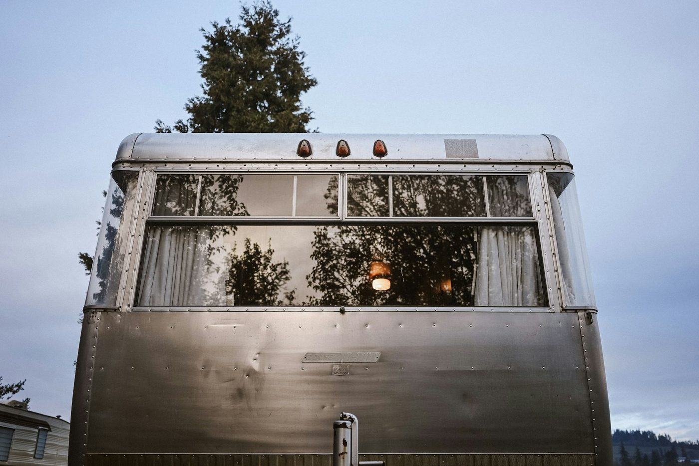A vintage silver travel trailer with a low-profile membrane roof, parked among trees